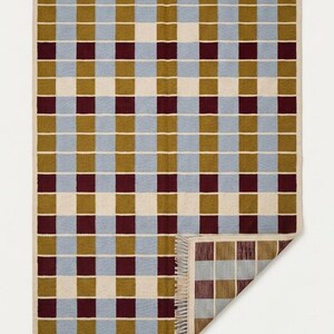 May include: A rectangular woven rug featuring a repeating grid pattern of light blue, olive green, cream, and deep burgundy squares. The rug has a fringed edge along the top and bottom, with one corner folded over to reveal the reverse side's texture.