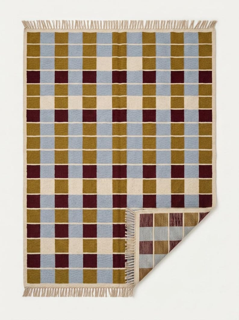 May include: A rectangular woven rug featuring a repeating grid pattern of light blue, olive green, cream, and deep burgundy squares. The rug has a fringed edge along the top and bottom, with one corner folded over to reveal the reverse side's texture.