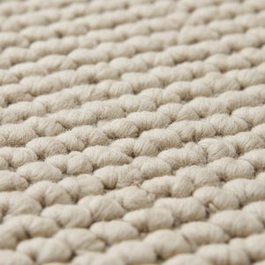 May include: Close-up of a beige, textured rug. The rug's surface is made up of tightly packed, rounded loops, creating a soft, tactile appearance. The loops are arranged in neat rows, giving the rug a uniform, yet organic, look.