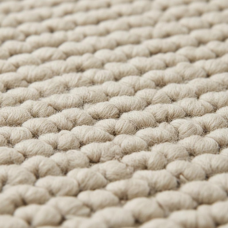 May include: Close-up of a beige, textured rug. The rug's surface is made up of tightly packed, rounded loops, creating a soft, tactile appearance. The loops are arranged in neat rows, giving the rug a uniform, yet organic, look.