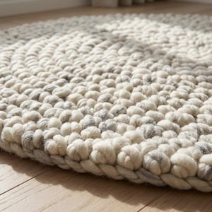 Puede incluir: Primer plano de una alfombra redonda y texturizada con borde trenzado. La alfombra est&aacute; hecha de lana blanquecina y gris, con un patr&oacute;n de c&iacute;rculos conc&eacute;ntricos. La alfombra est&aacute; sobre un suelo de madera clara.