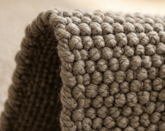 May include: Close-up of a textured, grey rug with a looped, bobble-like design. The rug's edge is visible, showing the thickness and construction of the woven material. The neutral colour and tactile surface suggest a cosy, home decor item.