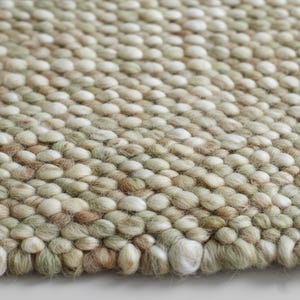 Pode incluir: Close-up de um tapete texturizado com um padrão de pequenos elementos arredondados e em relevo. O tapete apresenta uma mistura de cores neutras, incluindo tons de bege, verde claro e castanho. O design cria um efeito tátil tridimensional, adequado para decoração de interiores.