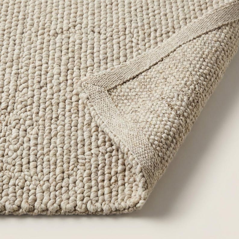 May include: A close-up of a beige, textured rug with a rolled-up corner. The rug has a looped pile and a woven border. The neutral color and texture suggest a cozy, natural aesthetic, suitable for home decor.