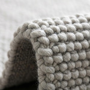 Peut inclure: Gros plan sur un tapis gris texturé avec un motif bouclé. Les fibres du tapis sont tissées serrées, créant un aspect doux et moelleux. La couleur neutre et la texture tactile suggèrent un élément de décoration intérieure confortable et moderne.