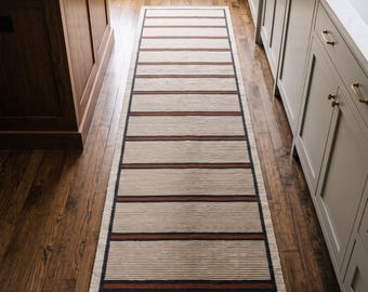 May include: A long rectangular runner rug featuring a cream-colored base with a repeating pattern of thin horizontal lines. The design includes wider, dark-colored stripes accented by thin, rust-colored bands that run across the width of the rug.