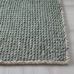 May include: Close-up of a textured area rug with a repeating pattern of rounded, woven fibers. The rug features a blend of light blue, gray, and white colors, with a white border. The rug is placed on a light-colored wooden floor.