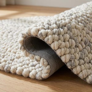 Puede incluir: Una alfombra enrollada y texturizada con una combinaci&oacute;n de colores crema y gris. La alfombra presenta un dise&ntilde;o en bucle, similar a una burbuja, creando un efecto tridimensional. La parte inferior de la alfombra es de color gris s&oacute;lido, sobre un suelo de madera.