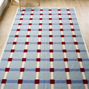 Könnte beinhalten: Ein rechteckiger Teppich mit hellblauem Hintergrund und einem Gittermuster aus sich kreuzenden weißen vertikalen und horizontalen Linien, das an jedem Schnittpunkt durch kleine, durchgehend rote Quadrate auf der gesamten Fläche akzentuiert wird.