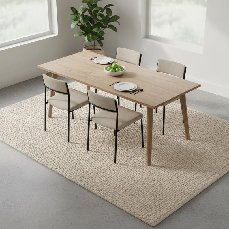 May include: A light wood dining table with four chairs, set on a cream-colored rug. The chairs have black metal frames and beige seats and backs. A bowl of green apples and place settings are on the table. A potted plant is in the background.