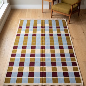 Pode incluir: Um tapete retangular com um padrão xadrez repetitivo em tons de azul claro, amarelo mostarda, creme e bordô escuro. O tapete tem uma textura tecida e é finalizado com franjas curtas e atadas nas duas extremidades mais curtas.