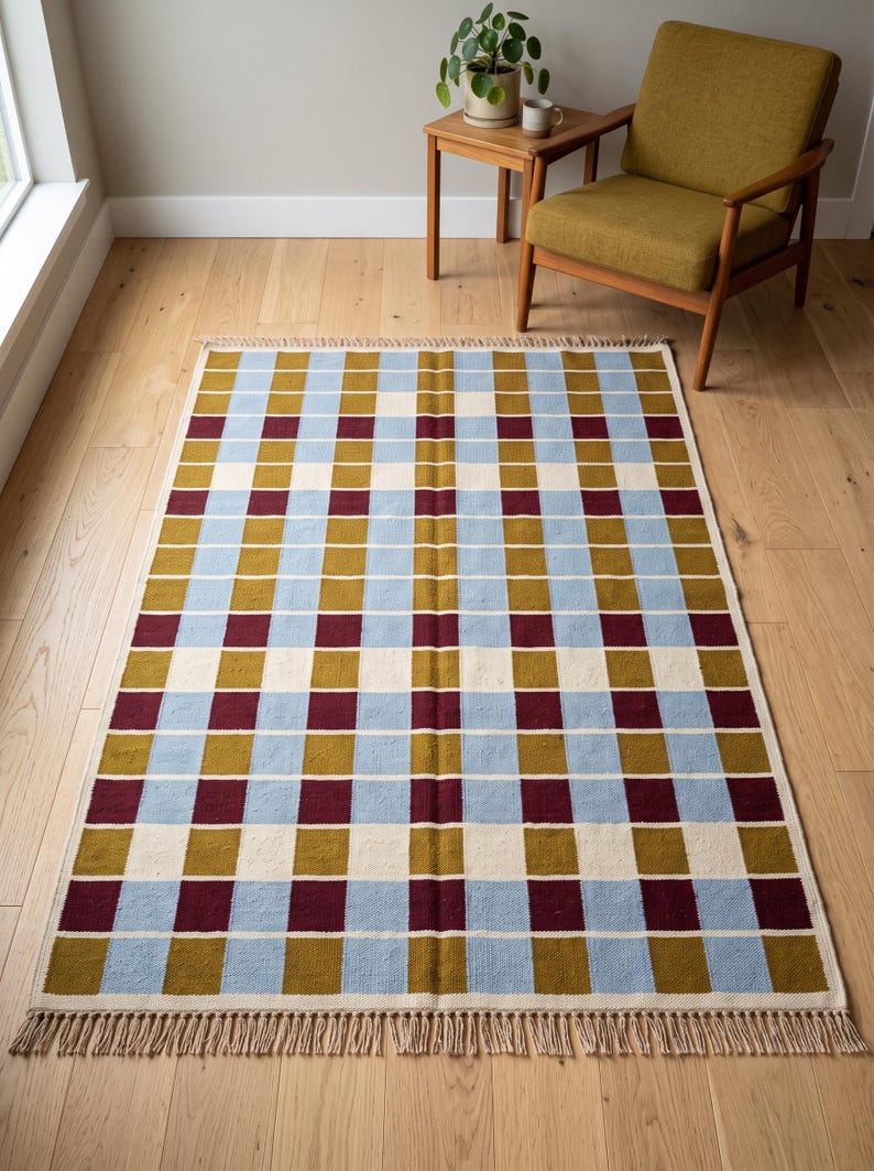 Handwoven flatweave checkered area rug in mustard, dusty blue and burgundy on a light wood floor beside a mid century mustard armchair in a bright modern living room.