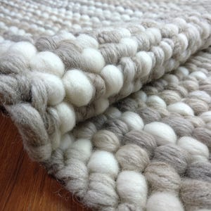 May include: Close-up of a textured rug with a repeating pattern of white, grey, and tan wool balls. The rug is folded over, revealing the underside. The handmade rug has a soft, plush appearance.
