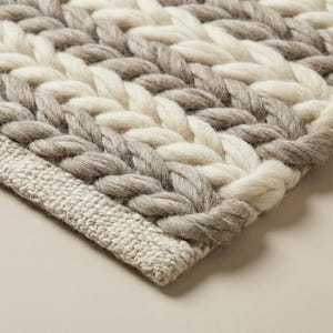 以下が含まれることがあります： 編み込みデザインの織りラグのクローズアップ。ラグは、ライトグレー、オフホワイト、ミディアムグレーの糸が交互に並んでいます。ラグの端には、ライトベージュ色のテクスチャードボーダーがあります。ラグは太い糸でできています。