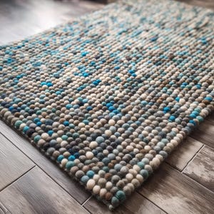 May include: A rectangular rug with a textured surface made of small, round, wool-like balls. The rug features a gradient of colours, including shades of blue, grey, and white. The rug is placed on a wooden floor.