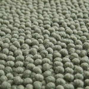 May include: Close-up of a sage green rug with a textured surface. The rug is covered in small, rounded tufts, creating a dense, tactile appearance. The colour is a muted, earthy green, providing a soft, natural aesthetic.