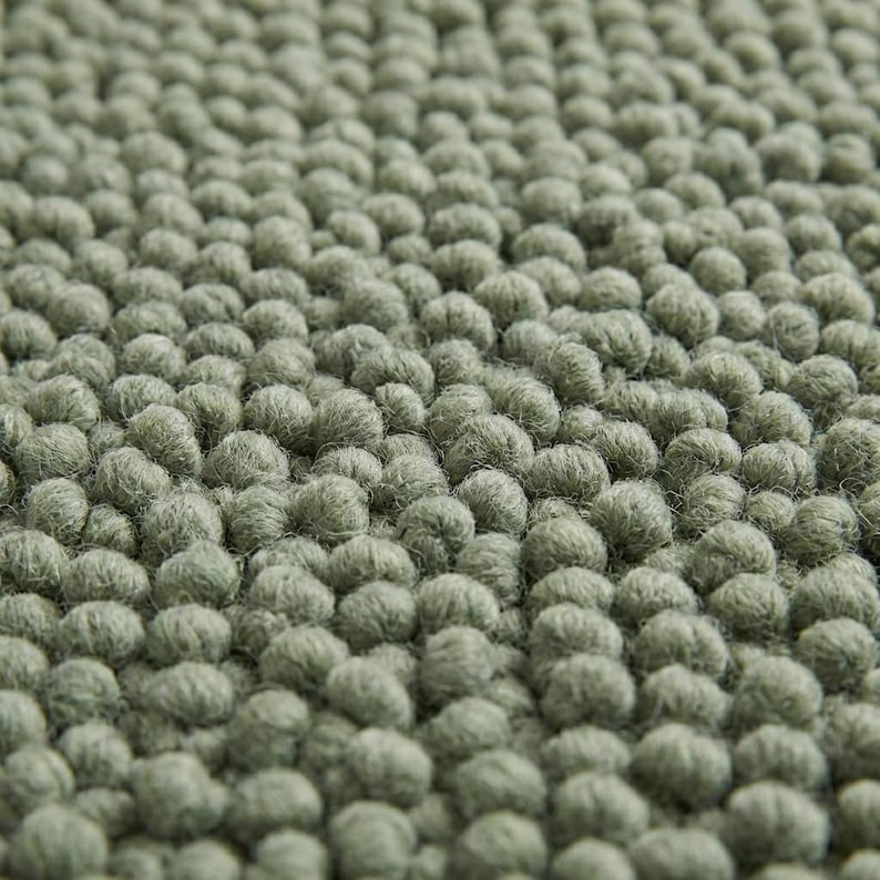 May include: Close-up of a sage green rug with a textured surface. The rug is covered in small, rounded tufts, creating a dense, tactile appearance. The colour is a muted, earthy green, providing a soft, natural aesthetic.