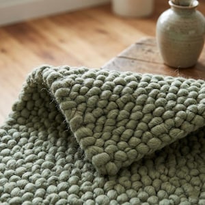 May include: A close-up shot of a sage green textured rug with a bubble-like pattern. The rug is folded over, showing its thick, plush texture. A light-coloured ceramic vase sits on a wooden table in the background.