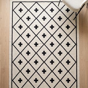 Könnte beinhalten: Ein rechteckiger Teppich mit cremefarbener Basis und einem sich wiederholenden schwarzen Rautenmuster. Jede Raute enthält ein zentriertes schwarzes Kreuzsymbol. Der Teppich hat einen durchgehenden schwarzen Rand, der das Design einrahmt.