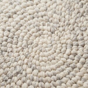 Puede incluir: Primer plano de una alfombra color crema con un dise&ntilde;o en espiral. La alfombra est&aacute; hecha de un material texturizado con peque&ntilde;os mechones redondeados en tonos crema y gris. El dise&ntilde;o crea un patr&oacute;n circular.