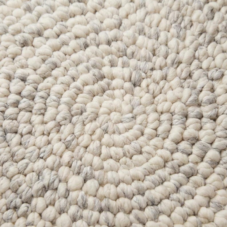 Puede incluir: Primer plano de una alfombra color crema con un dise&ntilde;o en espiral. La alfombra est&aacute; hecha de un material texturizado con peque&ntilde;os mechones redondeados en tonos crema y gris. El dise&ntilde;o crea un patr&oacute;n circular.
