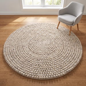 May include: A large, circular area rug constructed from numerous small, light beige and off-white felted wool balls arranged in a concentric spiral pattern. The rug features a textured, spherical surface and sits flat on a wooden floor.