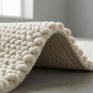 May include: Close-up of a cream-coloured, textured rug with a looped, bobble design. The rug's edge is visible, revealing a woven backing. The soft, neutral tones and tactile surface suggest a cosy, inviting home decor item.
