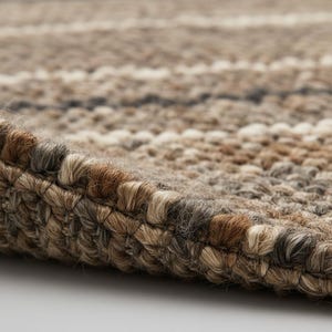 May include: Close-up of a textured rug with a woven design. The rug features a mix of neutral colors, including shades of brown, beige, and gray, creating a natural and earthy aesthetic. The edges of the rug are visible, showing the detailed craftsmanship.