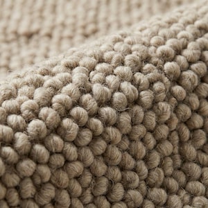 Puede incluir: Primer plano de una alfombra beige texturizada. La superficie de la alfombra está cubierta de pequeños bucles nudosos y redondeados, creando un patrón táctil y visualmente interesante. El color neutro y la textura densa sugieren un elemento de decoración del hogar acogedor y cómodo.