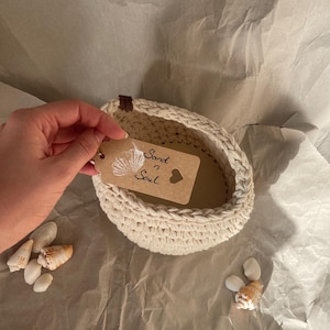 以下が含まれることがあります： クリーム色の、かぎ針編みのバスケット。茶色の革のアクセントが付いています。「Sand & Soul」とハートの文字が入ったタグが付いています。バスケットの周りには、いくつかの貝殻が散らばっています。