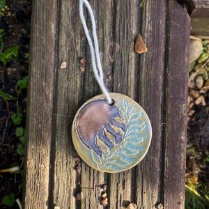 May include: A circular ceramic pendant with a pale blue cord. The pendant features a brown half-circle and a light blue half with a leaf design. It rests on a weathered wooden surface.