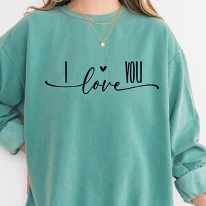 Puede incluir: Una sudadera de color verde azulado con la frase "I love you" en escritura negra. La sudadera tiene mangas largas y un ajuste relajado. El texto está centrado en la parte delantera de la prenda. Las palabras están conectadas por una línea curva.