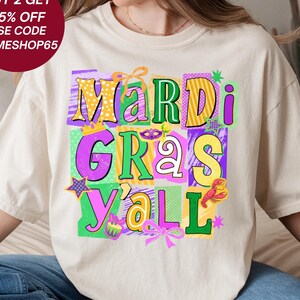 May include: A cream-coloured t-shirt with a colourful "Mardi Gras Y'all" graphic. The design features the text in yellow, green, purple, and pink, with festive icons like a crown, stars, and a crawfish. The words "Prime Digital Shop" are printed at the bottom.