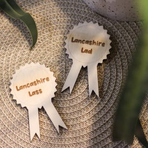 Könnte beinhalten: Zwei Holzrosetten mit Bandenden, auf denen die Worte "Lancashire Lass" und "Lancashire Lad" in brauner Schrift eingraviert sind. Sie liegen auf einer beigefarbenen, gewebten Oberfläche, mit Grün im Hintergrund.