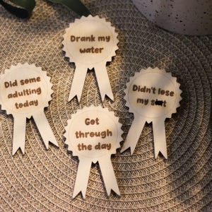 Könnte beinhalten: Vier hölzerne Auszeichnungen mit Schleifen. Jede Auszeichnung hat einen anderen Satz: "Drank my water", "Did some adulting today", "Didn't lose my sh*t" und "Got through the day". Die Auszeichnungen sind hellbraun und liegen auf einer strukturierten Oberfläche.