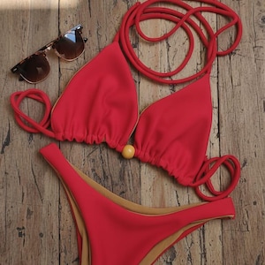 Puede incluir: Un conjunto de bikini rojo con top triangular y braguita de cintura alta. El top tiene un diseño de cordones y una pequeña cuenta amarilla. El bikini se muestra sobre una superficie de madera, con gafas de sol.