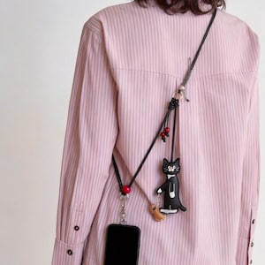 May include: A black phone case with a black strap, featuring a black cat charm wearing a tuxedo and holding a small fish. The strap has red beads and a silver clasp. The phone case is hanging on a pink striped shirt.
