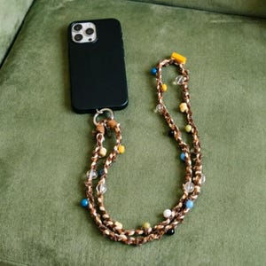 May include: A black smartphone case with a decorative lanyard. The lanyard is made of brown braided cord, adorned with colorful beads in yellow, blue, clear, and white. The phone case is attached to the lanyard with a silver-colored ring.