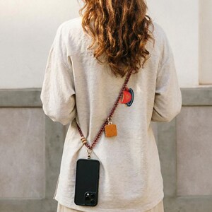 Puede incluir: Un teléfono inteligente negro con una funda cuelga de una correa de cuerda burdeos y marrón. La correa presenta cuentas de madera y una pequeña etiqueta cuadrada de cuero. Un diseño circular colorido es visible en la parte superior de la espalda de la camisa.