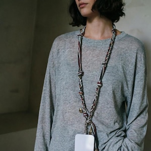 Puede incluir: Una funda blanca para smartphone con un cordón trenzado. El cordón es multicolor con detalles en rojo, blanco y negro, y una pequeña campana dorada. La funda del teléfono cuelga del cordón.