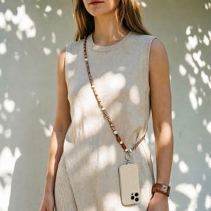 Può includere: Un top beige senza maniche abbinato a una lunga tracolla per telefono con piccole perline marroni, bianche e rosse. Il cinturino si collega a una custodia per smartphone beige tinta unita tramite un anello in metallo argentato all'altezza dell'anca.