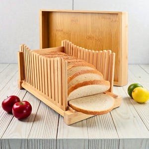 May include: A bamboo bread slicer with a loaf of sliced bread. The slicer has vertical slots to guide the knife and a base to catch crumbs. A wooden tray is behind the slicer. Two red apples, a lime, and a lemon are on the table.
