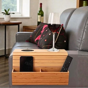 May include: A bamboo sofa armrest table with a built-in cup holder, two compartments, and a phone slot. A wine glass with red wine sits on the table. A remote control is in one compartment.
