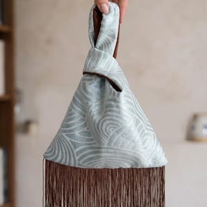 May include: A light blue fabric bag with a textured wave pattern and a brown interior lining. The bag features a unique knot handle design and a long, straight brown fringe trim along the bottom edge, creating a distinct triangular silhouette.