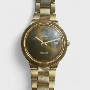 May include: A gold-toned Omega De Ville Dynamic wristwatch. The watch features a dark green face with gold hands and hour markers, and a date display at the 3 o'clock position. The watch band is also gold-toned and has a classic link design. The watch is set against a neutral background.