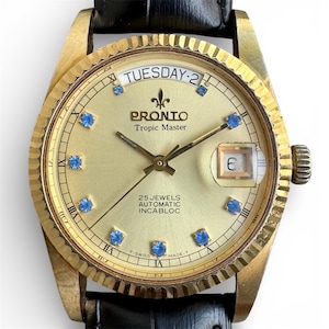 May include: A gold-toned Prono Tropic Master watch with a black leather band. The watch face displays the day and date, with blue gemstone hour markers. The text "TUESDAY-2" is visible at the top, and "25 JEWELS AUTOMATIC INCABLOC" is at the bottom.