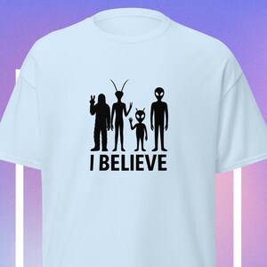May include: Light blue t-shirt with a black graphic of four alien figures and the text "I BELIEVE". The figures include a sasquatch-like silhouette, a tall alien with antennae, a small alien, and a classic alien.
