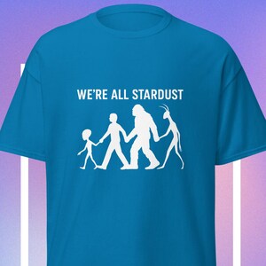 May include: A turquoise t-shirt with the text "WE'RE ALL STARDUST" above a white graphic of a child, a person, a Bigfoot, and an alien holding hands. The shirt is a short-sleeved, crew neck style.