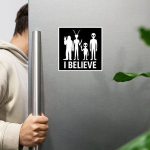 May include: A white magnet with a black square background featuring four alien figures and the text "I BELIEVE" in white. The aliens are in various poses, with one giving a peace sign.