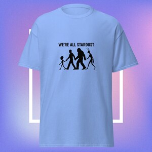 May include: Light blue t-shirt with the text "WE'RE ALL STARDUST" above a black graphic of a child, a person, a Bigfoot, and an alien holding hands. The shirt has short sleeves and a crew neck.